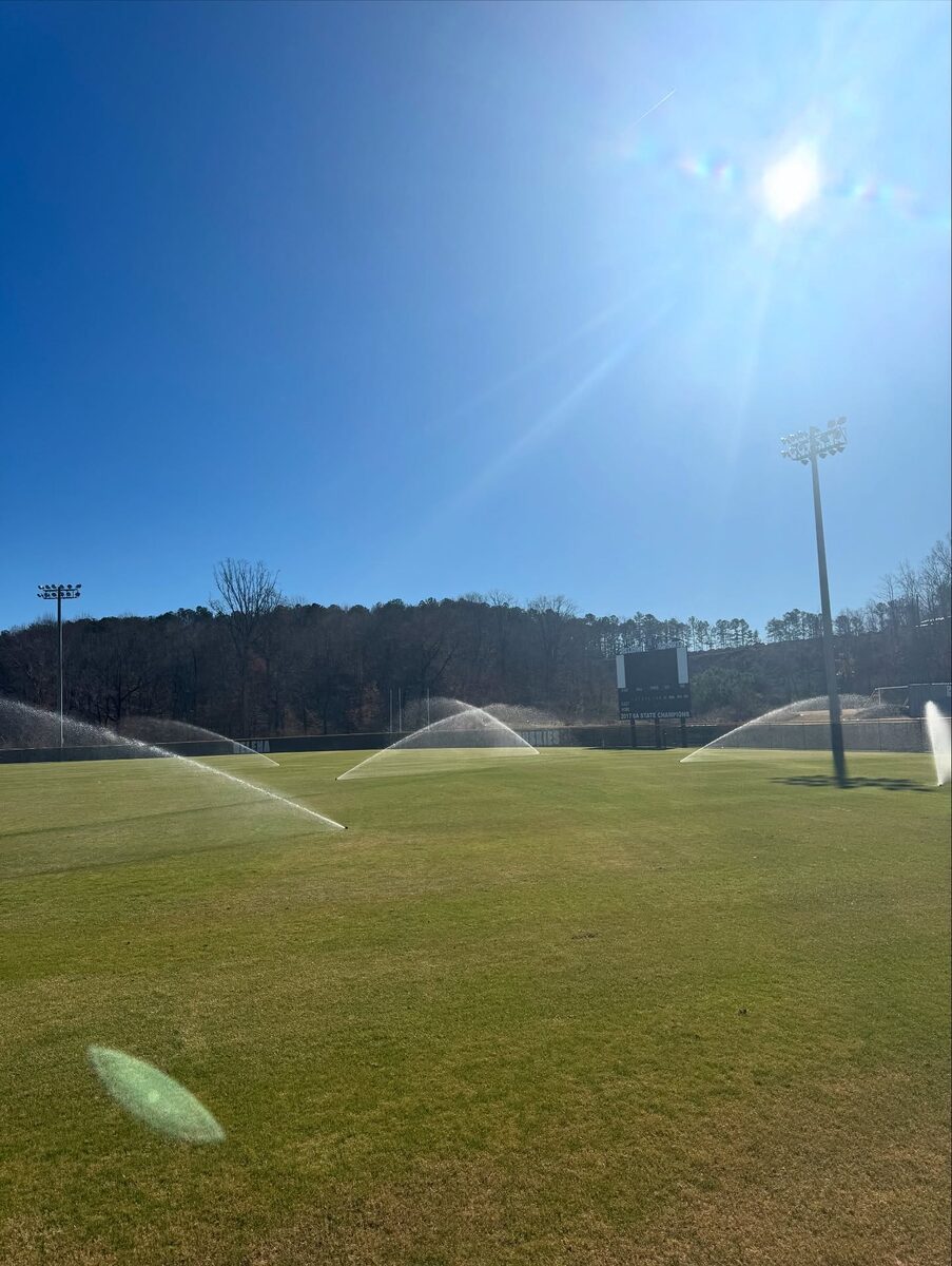 Irrigation heads running across a large sports field under a bright sky.