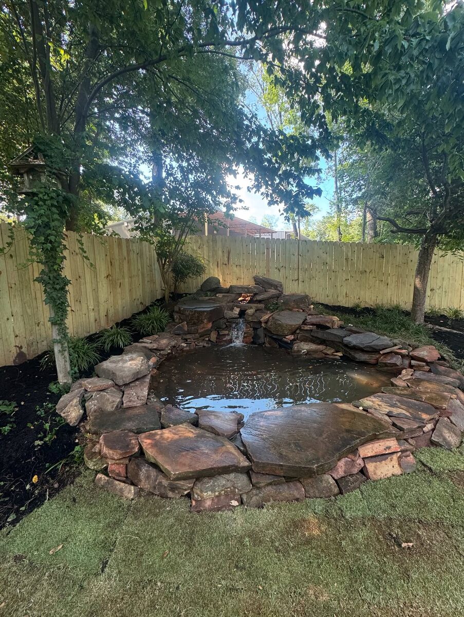 A stone-edged garden pond set into a lawn against a wooden privacy fence.