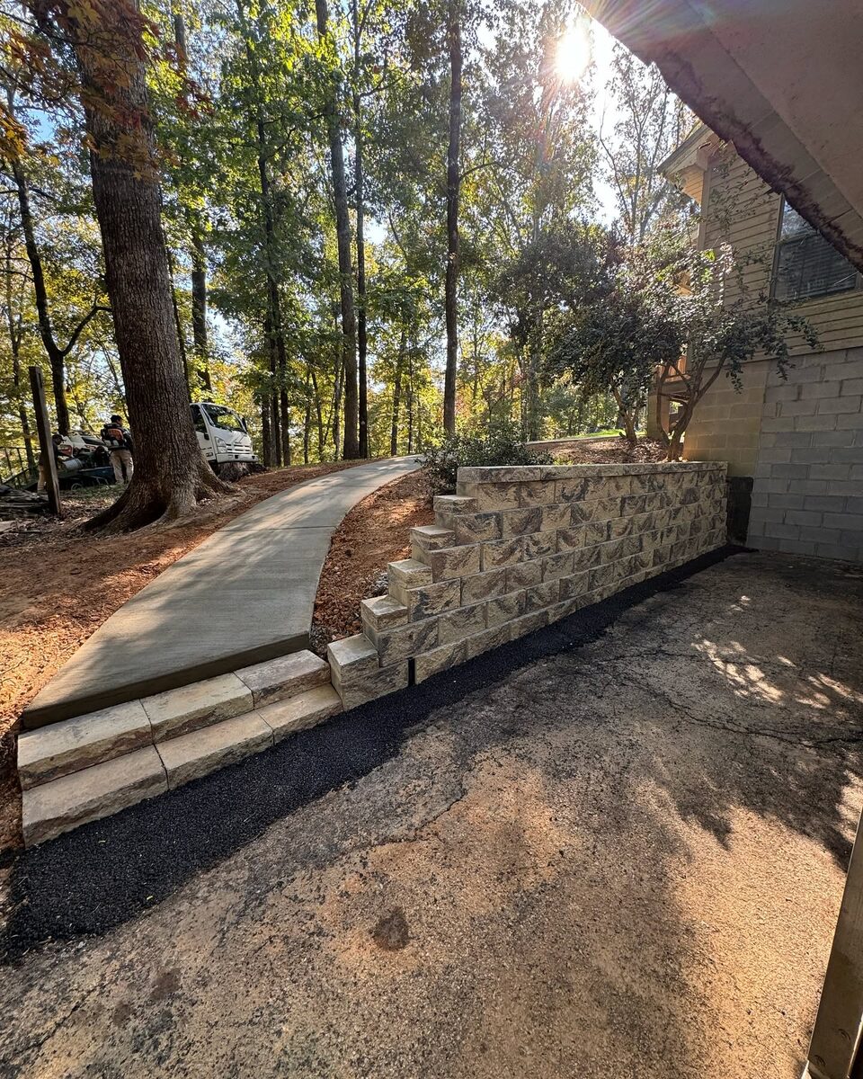 A stacked stone retaining wall running beside a new concrete walkway on a wooded slope.